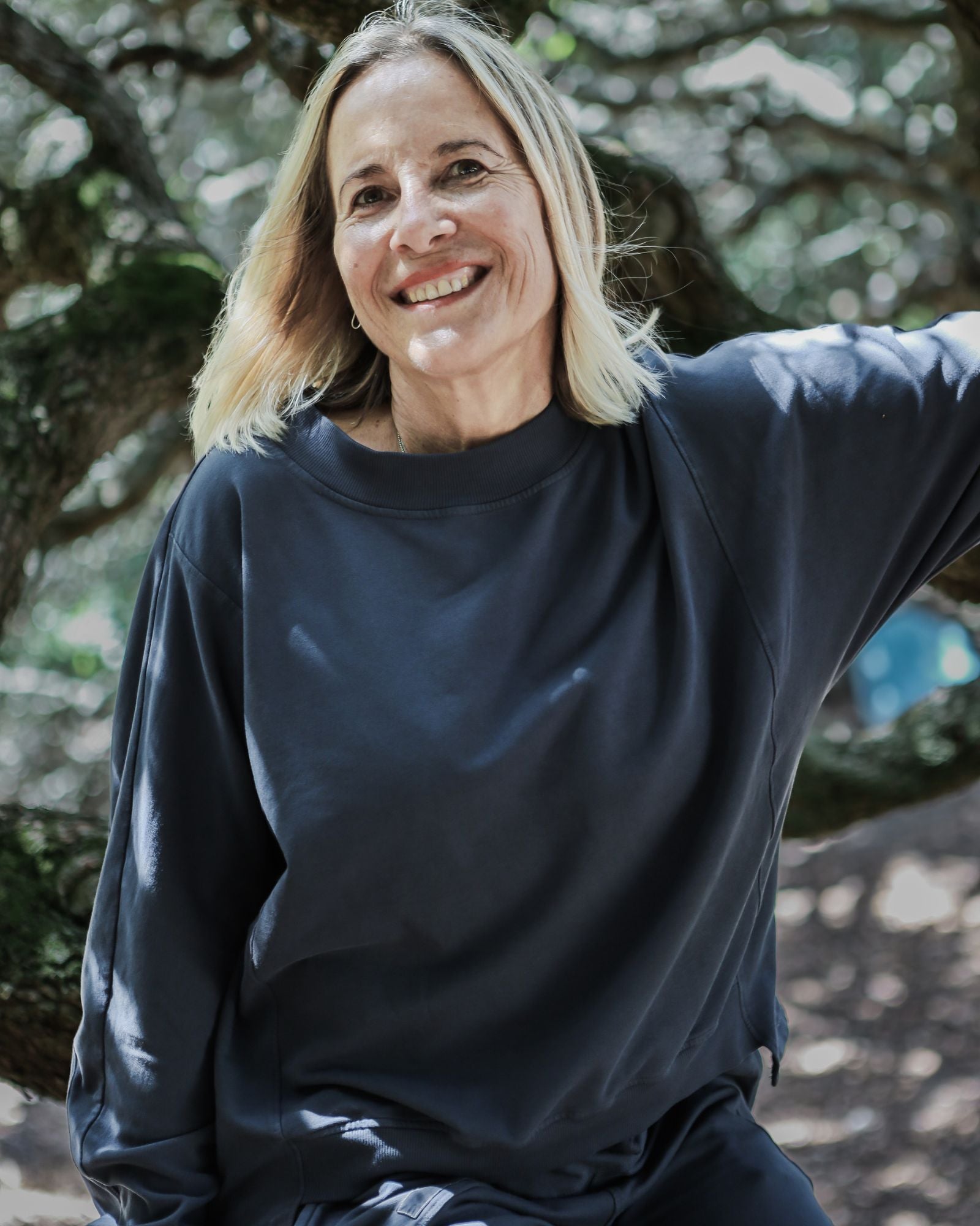 Woman wearing a charcoal color sweatshirt sitting outdoors with trees in the background